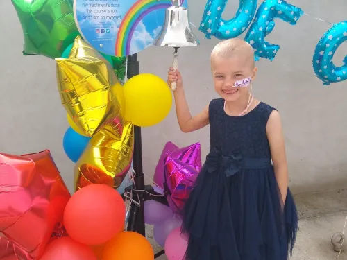 A young girl with a bald head and a nasogastric tube is ringing the End of Treatment Bell. She is wearing a navy party dress, surrounded by colourful balloons and decorations.