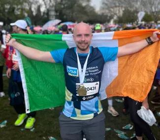 A man stands with a flag held behind his back, wearing a CCLG: The Children and Young People's Cancer Association running t-shirt. A medal hangs around his neck, showcasing his achievement.