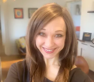 A close-up headshot of a smiling woman with brown hair wearing a brown shirt